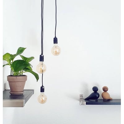 Nielsen Light Klyngependel K9 med 3 sorte tekstilledninger og dekorative pærer, monteret på væggen ved siden af en plante og fuglefigurer.