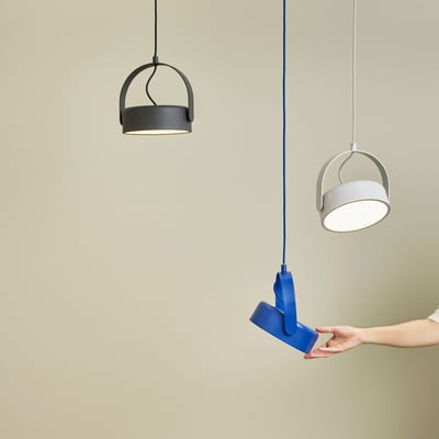 En hånd holder Hübsch Stage loftlampen i blå metal ved siden af to andre Stage lamper i grå og hvid.