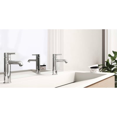 To Oras Optima Style krom håndvaskarmaturer står på en hvid vask, med et spejl og en grøn plante i baggrunden.