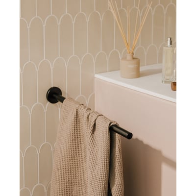 Cosmic håndklædeholder i mat sort med en lys beige vaffelhåndklæde hængende. Holderen sidder op ad en væg med beige buede fliser.