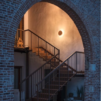 SLV Mana Base Phase væglampe i antracit grå monteret på en væg ved en trappe, lyser varmt.