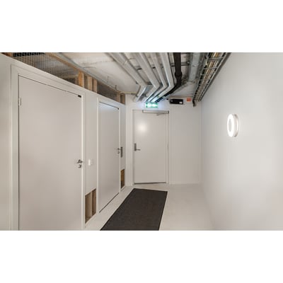 SG Armaturen Etne væg-/loftarmatur i hvidt polycarbonat med 4000K lys på en hvid væg i en gang med døre og rør i loftet.