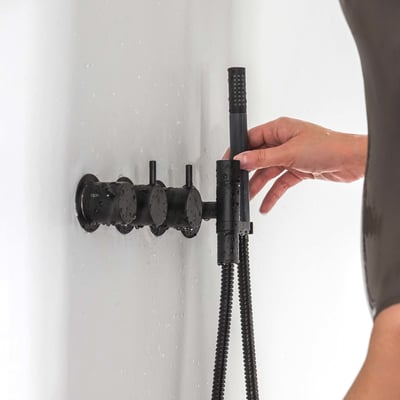 Vola indbygningstermostat i matsort med tre greb og håndbruser, der betjenes af en hånd på en hvid flisevæg med vandsprøjt.