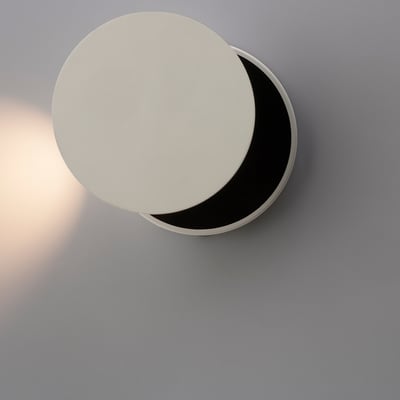 ANTIDARK Dot W120 LED sengelampe i hvid aluminium med en diameter på 120 mm, monteret på en grå væg og oplyst nedad.