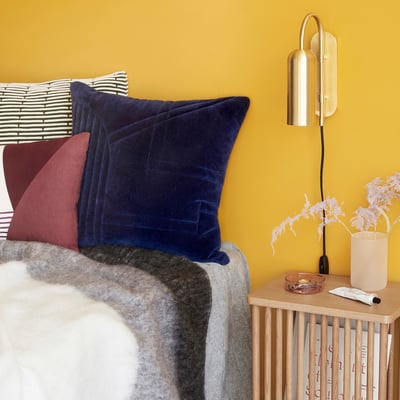 Hübsch Zenith væglampe i messingfarve, monteret over en seng med gule vægge og blå puder.