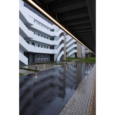 Moderne bygning med udendørs belysning, der reflekterer på våde fliser og asfaltsti.