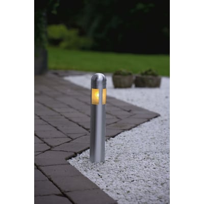 En enkelt tændt Konstsmide Amalfi mini LED bedlampe står på en hvid stenbelagt overflade med græsplæne i baggrunden.