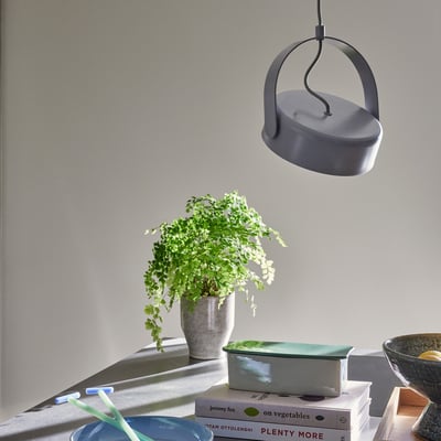 Detaljebillede af den grå Hübsch Stage loftlampe, der hænger over en potteplante og bøger på en bordplade.