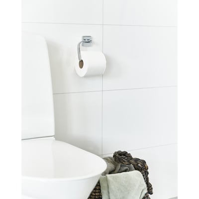 Smedbo toiletpapirholder i poleret krom monteret på et hvidt, flisebelagt badeværelsesvæg med en toiletpapirrulle påsat.