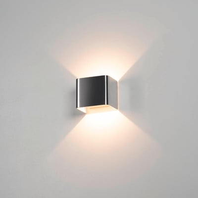 SLV MANA lampeskærm monteret på en grå væg, oplyst med et varmt lys der skaber lysstriber opad og nedad.