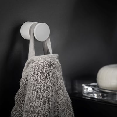 Cosmic håndklædekrog i mat hvid, vist monteret på en mørk væg med et gråt håndklæde hængende på.