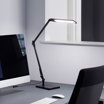 Nielsen Light arkitektlampe i sort, med justerbar arm og lampehoved der lyser, placeret på et kontorbord ved siden af en computerskærm.