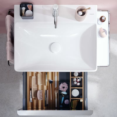 Hansgrohe IntraStoris skuffeinddeler i natur eg, vist i en åben skuffe med diverse toiletsager og kosmetik arrangeret.