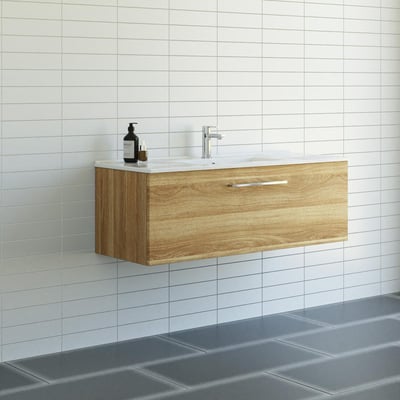Dansani Mido møbelpakke på 121 cm med varm eg-finish og en skuffe, monteret på en hvid væg i et moderne badeværelsesmiljø med fliser.