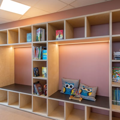 Indbygget reol med lyserøde vægge og åbne rum fyldt med bøger, legetøj og dekorative puder med uglemotiv.