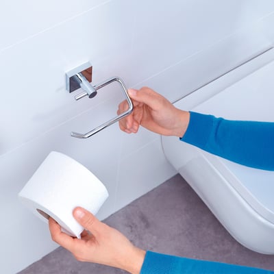 Tesa® Ekkro toiletpapirholder i krom monteres på hvid flisevæg af en person, som holder en toiletpapirsrulle.