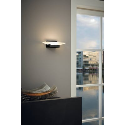 EGLO Metrass væglampe i sort aluminium med et 200mm langt, moderne design, monteret på en grå væg ved et vindue med udsigt til vand og bygninger.