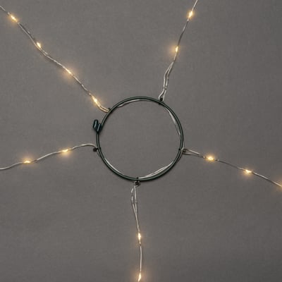 Konstsmide juletræskæde med 150 rav LED-lys arrangeret omkring en grøn metalring, vist på mørkegrå baggrund.