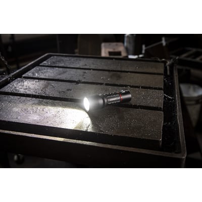 Coast XP11R lygte med lysende LED-pære, placeret på et rustent metalbord i et værkstedsmiljø.