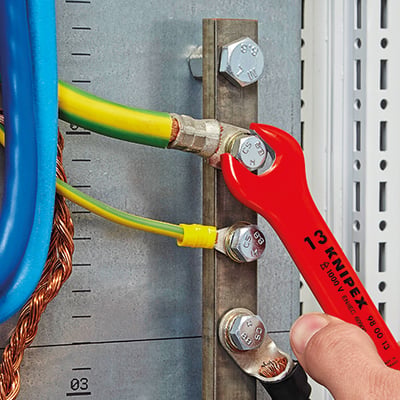 Hånd der bruger en rød KNIPEX L-AUS fastnøgle til at spænde en bolt på en elektrisk samleskinne med flere kabler tilsluttet.