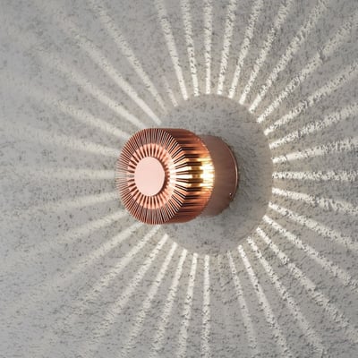 Konstsmide Monza væglampe i kobberfarvet aluminium på en hvid væg, der udsender et dekorativt lysmønster.