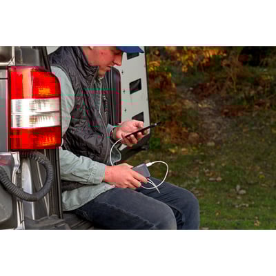 NEBO 5000 mAh powerbank oplader en smartphone for en mand, der sidder ved bagenden af en bil i naturen.