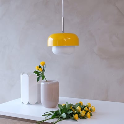 Dyberg Larsen HAIPOT pendel i gul, monteret over et bord med gul blomsterdekoration og lysegrå vase.