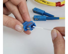 En hånd bruger en 2,5 mm fiberoptisk rensesvab med skum til at rengøre et blåt SC-fiberstik, mens andre stik ligger klar.
