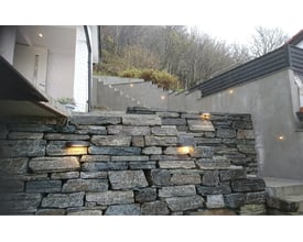 Vægtarmatur Curve Direct fra SG Armaturen monteret på en stenmur ved en trappe, giver varmt hvidt lys udendørs.