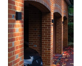 Astro 7202 Elis Twin sort væglampe monteret på en murstensvæg ved en garageindkørsel med efterårsblade på jorden.