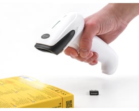 Delock 1D/2D barcode scanner i hånden, klar til at scanne en gul produktæske og viser en lille USB dongle på bordet.