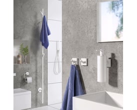 Smedbo House toiletpapirholder i mat hvid monteret på en grå badeværelsesvæg, ved siden af en hvid vask og sæbedispenser.