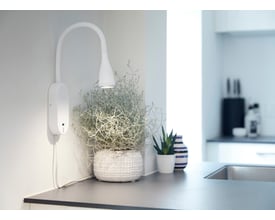 Nielsen Light Eye væglampe i hvid metal med fleksibel hals og USB-port, monteret ved siden af planter og en vase.
