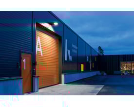 SG Armaturen Langfoss Mini projektør oplyser en stor orange garageport med bogstavet A og en mindre orange dør med tallet 1. Bygningen er mørk med blålig aftenhimmel.