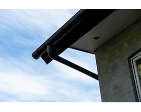 Plastmo sort tagrendesystem monteret på husmur med blå himmel i baggrunden, viser endebund og nedløbsrør.