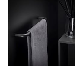 Cosmic håndklædeholder i mat sort, 40 cm bred, med et gråt håndklæde hængende på. Holderen er monteret på en sort væg, og til højre ses en lille grå hylde med en glasvase og sorte pinde.