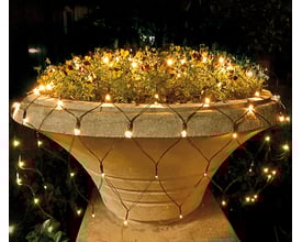 Veli Line udendørs LED lysnet med rav guld lys, 300x400 cm, monteret omkring en stor stenopsats med blomster i mørket.