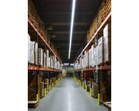 Langt LEDVANCE TruSys Flex armatur monteret i et lager med reoler fyldt med varer, og oplyser midtergangen med et bredt hvidt lys.