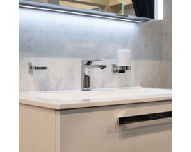 Smedbo House sæbedispenser i poleret krom med materet glas monteret på badeværelsesvæg ved siden af en vandhane og et glas.