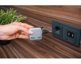 En hånd holder Verbatim mini GaN-opladeren på 65 W ved en stikkontakt; displayet vender væk.