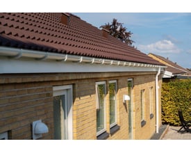 Hvid Plastmo brøndkrave monteret på et hus med gule mursten og brunt tegltag.