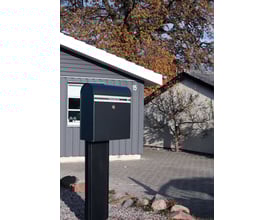 Assa Abloy Allux KS 200S postkasse i sort stål monteret på sokkel i forhave med efterårsfarver og hus i baggrunden.