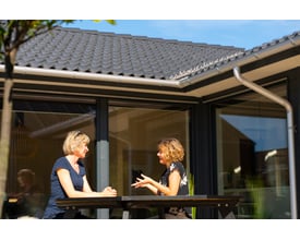 To kvinder taler sammen på en terrasse, mens Plastmo tagrenden i aluzink ses langs husets tagkant.