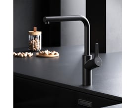AM.PM X-Joy S køkkenarmatur i mat sort messing med svingbar L-tud og etgrebsbetjening, monteret på sort bordplade med nødder i baggrunden.