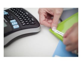 Dymo LabelManager 210D labelprinter i sort og sølv, hvor en hånd påsætter en label på en grøn mappe.