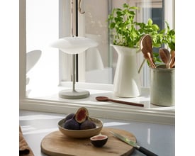 Halo Design Baroni Move bordlampe i varm grå med hvid skærm, placeret på en vindueskarm med grønne planter og frugt.