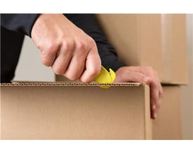 Hånd bruger en gul Klever KCJ-1 sikkerhedsskærer til at åbne en papkasse, hvilket viser dens anvendelse.