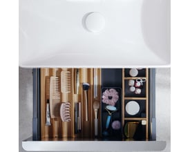 Hansgrohe IntraStoris skuffeinddeler i natur eg med makeup og toiletsager, sat i en åben skuffe med vask i baggrunden.