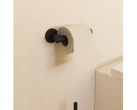 Cosmic Architect S+ toiletpapirholder i mat sort, monteret på en lys væg med en rulle toiletpapir påsat.