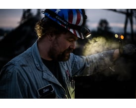 Mand med pandelampe Coast XPH34R på hovedet, oplyser arbejdsemne med kraftigt lys i mørke omgivelser.